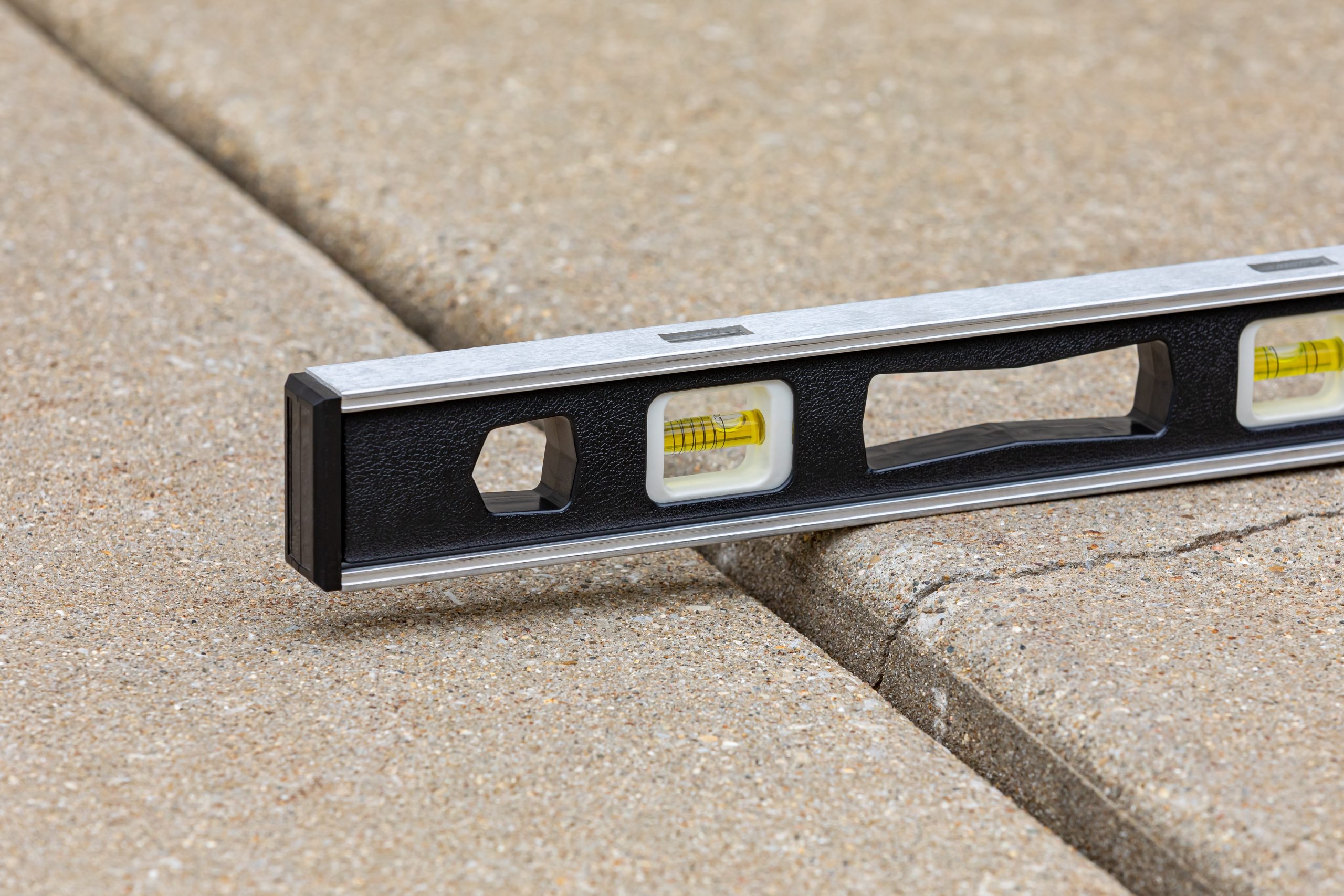 Uneven concrete sidewalk with raised slab highlighted by a level tool showing misalignment – trip hazard and foundation repair concept.
